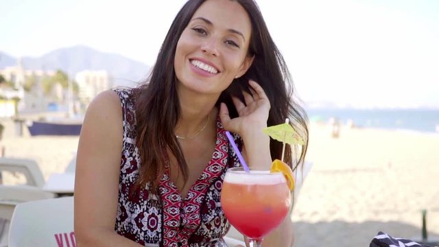 Happy friendly young woman drinking a tropical cocktail as she sits at an open air beachfront restaurant relaxing on her summer vacation
