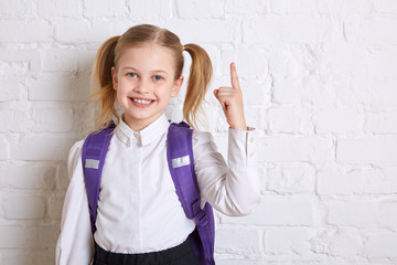 Cute smiling schoolgirl in uniform standing on light  background and showing  thumb up. Copy space