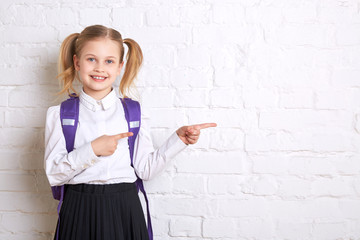 Cute smiling schoolgirl in uniform standing on light  background and showing  thumbs to the side. Copy space