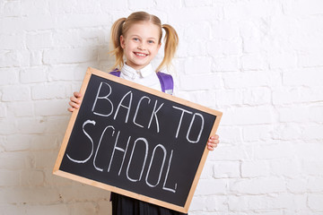 Cute smiling schoolgirl in uniform standing with blackboard and smiling on light  background. Back to school.