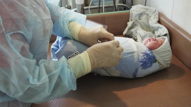 Nurse in gloves tie up nameplate on swaddled newborn baby after birth on table. Diapers, blanket. Maternity hospital.