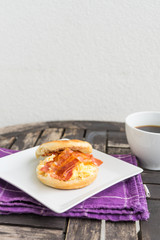 Salt muffin with scrambled eggs, bacon and cheese on white plate with dark coffee in white mug lying on wooden background. Unhealthy breakfast with bacon, eggs, pastry and coffee on purple dishtowel. 