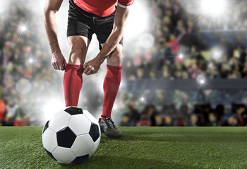 football player with ball wearing black shoes adjusting his red sock standing on stadium pitch