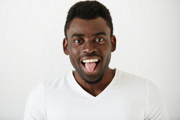 Close up portrait of African American student boy wearing white T-shirt making funny face, looking and showing tongue at the camera after classes at university. Attractive young male model having fun