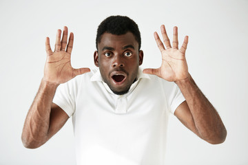 Portrait of stunned young African American man wearing white blank T-shirt looking at the camera in shock, surprised with some unexpected news, gesturing with hands. Human expressions and emotions