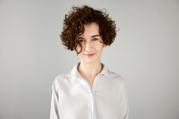 Close up portrait of fashionable hipster female with short curly hairstyle wearing white trendy shirt, having fun, looking at the camera with mysterious smile. Human face expressions and emotions