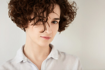 Isolated headshot of beautiful hipster girl with short curly hair wearing white trendy shirt looking at the camera. Portrait of young female with messy hairstyle relaxing at home. Lifestyle concept