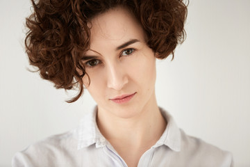 Close up shot of young brunette Caucasian woman looking with serious expression at the camera. Portrait of displeased or angry female office worker posing against white concrete wall background