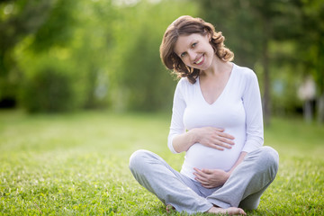 Fototapeta premium Smiling future mom sitting cross-legged on grass lawn