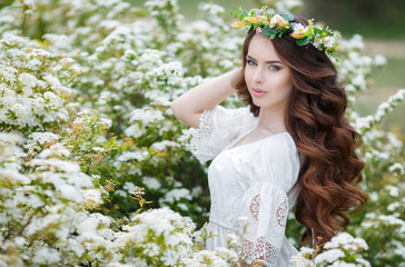 Obraz premium Portrait of a beautiful woman in a wreath of flowers,long curly red hair,gray eyes,a beautiful smile,dressed in a white summer dress posing near spring Bush of a blossoming white flowers