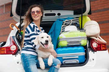 Beautiful young woman in sunglasses, wearing a t-shirt with white and dark blue stripes,blue jeans with holes in the knees,sits with his white dog near the white car, loaded with stuff