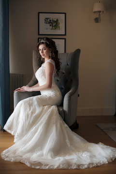 Beautiful young woman with brown eyes,professional makeup,brunette with beautiful wedding hairstyle and tiara,ears expensive earrings,dressed in a white wedding dress sitting on the armchair