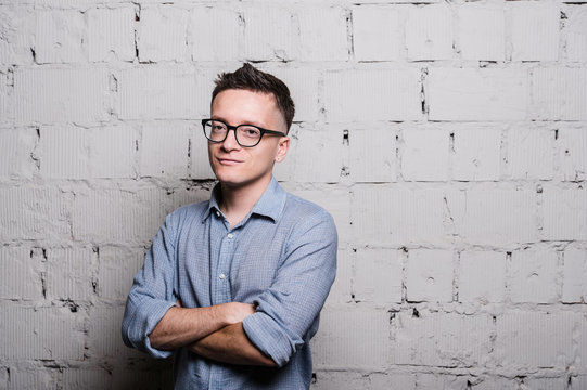 Portrait of handsome young man in jeans clothes and eyeglasses looking at camera smiling, standing against gray brick wall