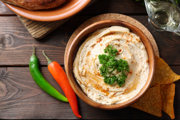 Wooden bowl of tasty hummus with chips and parsley on table