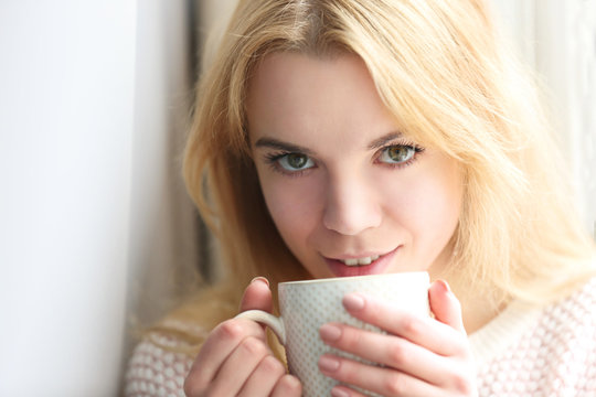 Blonde beautiful  girl sitting by the window with cup of tea or coffee in her hands