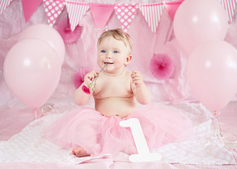 Obraz na płótnie Canvas Portrait of cute adorable Caucasian baby girl with blue eyes in pink tutu skirt celebrating her first birthday with gourmet cake and balloons looking away, cake smash first year concept