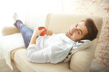 Naklejka premium Handsome man lying on sofa with red cup of coffee