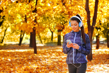 Young beautiful woman holding phone, listening to music with headphones in the autumn park