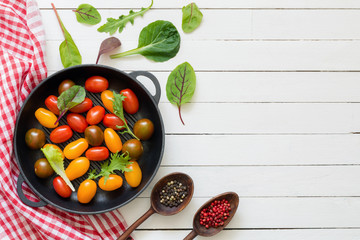Naklejka na ściany i meble Multi colored cherry tomatoes in pan, spices, kitchen textile and fresh green salad leaves on white wooden backdrop. Top view, flat lay food. Copy space for text