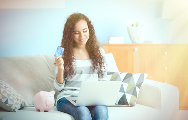 Beautiful young girl with credit card, piggy bank and laptop on couch
