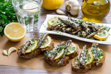Snack from sandwiches with sardines. In the foreground sandwiches with fresh sardines, in the background a plate of sardines, a glass of water, parsley, garlic, lemon, olive oil. Horizontal. Daylight.