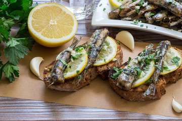 Snack from sandwiches with sardines. In the foreground sandwiches with fresh sardines, in the background a plate of sardines, a glass of water, parsley, garlic, lemon, olive oil. Horizontal. Daylight.