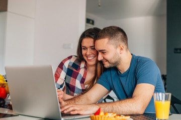 Young couple sitting in the kitchen. Man is using laptop. 