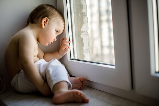 Adorable little kid boy sitting near window and looking on the street, indoors.