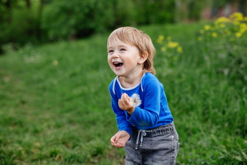 Happy smiling toddler  boy playing in a blooming garden on a meadow green grass during summer day holding flower dandelion