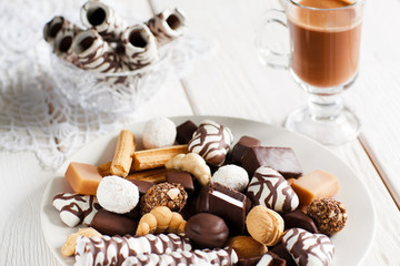 Delicious chocolate dessert on a white wooden table with cup of coffee cappuccino . Vase with cookies close-up on lace white napkin. Front view of chocolate candies, focus on foreground.