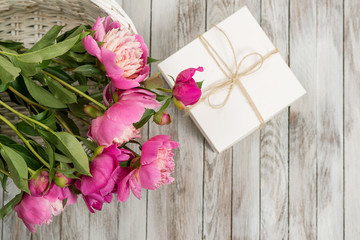 Fototapeta na wymiar Beautiful flowers peonies in basket with gift box on light wooden background. Top view.