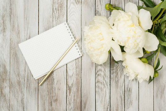 White peonies flowers with notebook and pencil on white painted wooden planks. Place for text. Top view.