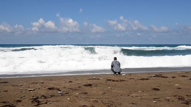 Funny fall. A man looks at the waves of the sea, the waves suddenly reach him, he tries to run away and falls. Sunny windy day in Cyprus. Back view