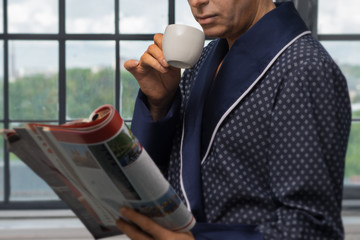 A man in a fashionable coat, sitting near the window, drinking coffee at breakfast and reading a...