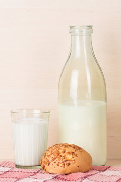 Old fashioned opened bottle of milk stands with faceted glass of milk and a bun on a red squared kitchen towel.