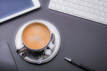 Gefüllte Kaffeetasse umrahmt von Tastatur, Smartphone, Tablet und Kugelschreiber