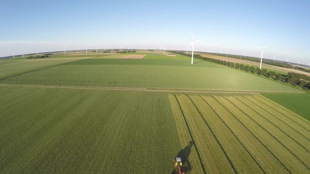 Aerial drone flying over a farmer in tractor working on grassland mixing mowing turning fresh grass in landscape with wind turbines windmills providing green energy renewable energy bird eye heli 4k