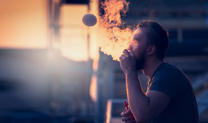 young man with beard on blurry background sunset sky, making soap bubbles with smoke inside with the aid of vape