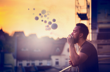 young man with beard on blurry background sunset sky, making soap bubbles with smoke inside with the aid of vape