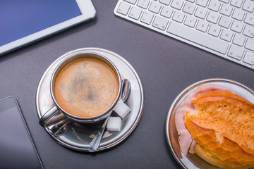 Gefüllte Kaffeetasse umrahmt von Tastatur, Smartphone, Tablet, Kugelschreiber und Frühstücksbrötchen