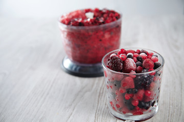 Mix of frozen berries in rox glass in front of unfocused blender pot with blended together berries and crashed ice cubes inside to make a healthy cold smoothie drink, top view on rustic table