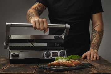 Man cooks healthy meal, prepares electronic home grill to roast two raw pieces of salmon decorated with mint leaf in white wine sause with spices and herbs presented on marble deck