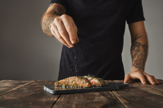 Man cooks healthy meal on rustic wooden table, salts two raw pieces of salmon in white wine sause with spices and herbs presented on marble deck prepared for grill 