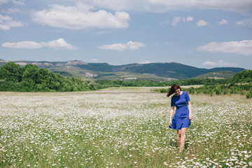 Fototapeta na wymiar Young beautiful woman in a blue dress enjoying chamomile field among mountains. Summer mood