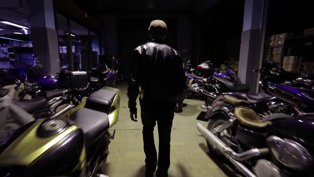 Young biker walks among motorcycles on a warehouse. Steadicam stabilized shot.