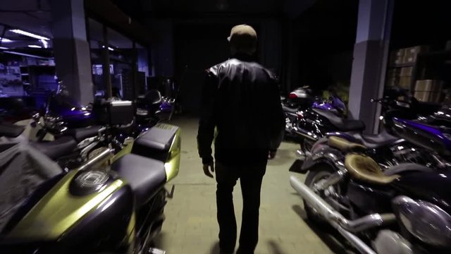 Young biker walks among motorcycles on a warehouse. Steadicam stabilized shot.