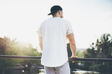 Photo Bearded Muscular Man Wearing White Blank t-shirt, snapback cap and shorts in summer time. Green City Garden Park Sunset Background. Back view. Horizontal Mockup