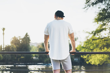 Photo Bearded Muscular Man Wearing White Blank t-shirt, snapback cap and shorts in summer vacation. Relaxing time near the lake. Green City Garden Park Sunset Background. Back view. Horizontal Mockup
