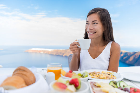 Asian Woman drinking hot drink at morning Breakfast table. Beautiful elegant lady having coffee cup on luxury hotel terrace with sea view at resort restaurant on Oia Santorini island, Greece.