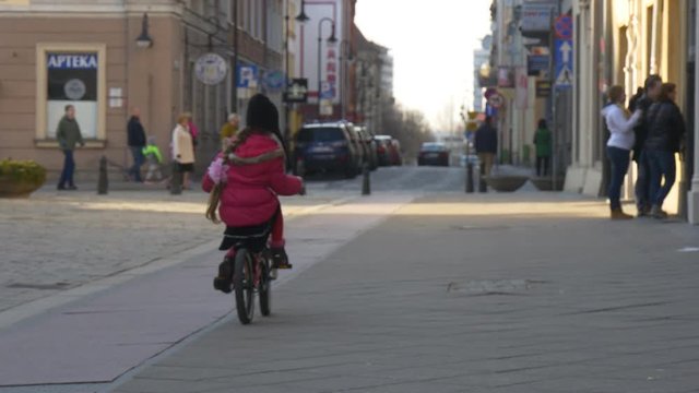 Little Girl is Riding the Bike Fast by City Square in Opole Poland Child is Riding Among Crowd of People Walking by a Street in Sunny Day Springtime